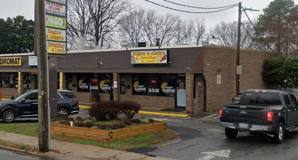 A wider shot showing the single-story commercial strip building housing “Bagner & Alexa’s Breakfast & Lunch” along with adjacent businesses like a laundromat. A roadside sign pole on the left displays the restaurant name, while a raised wooden planter box sits near the asphalt parking area. A black pickup truck is parked on the right side of the lot under an overcast sky.